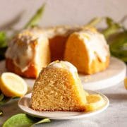Close-up of a slice of lemon Bundt cake with glaze on a white plate with a lemon slice, with the sliced cake in the background.