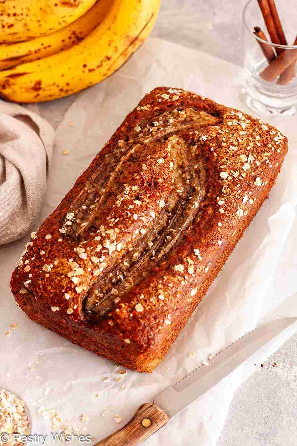 An overhead shot of a loaf of dairy-free banana bread on parchment paper, next to ripe bananas, a knife, some rolled oats and a glass filled with cinnamon sticks.