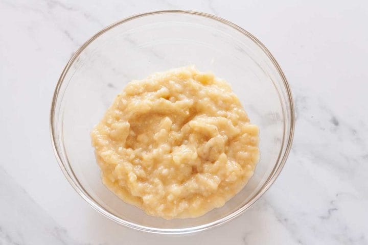 Mashed bananas in a glass bowl on a marbled white surface.