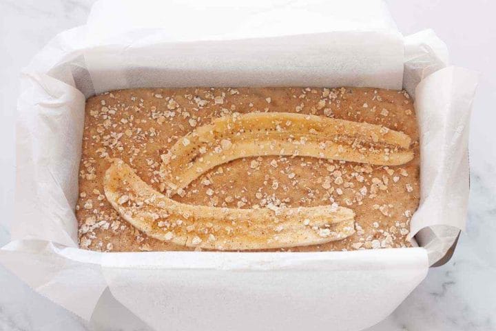Dairy-free banana bread batter in a lined loaf pan with rolled oats and turbinado sugar on top before baking.