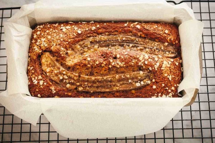 A baked loaf of dairy-free banana bread in a baking pan on a wire rack lined with parchment paper.