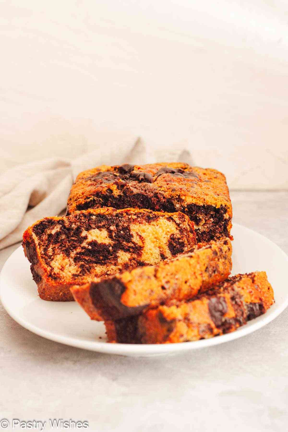 Three slices of marble loaf cake in front of a loaf cake on a white plate.