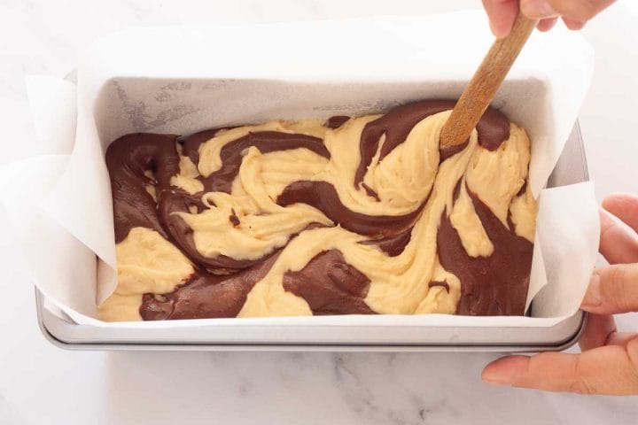 Swirling vanilla and chocolate cake batter with the back of a wooden spoon in a loaf pan lined with parchment paper.