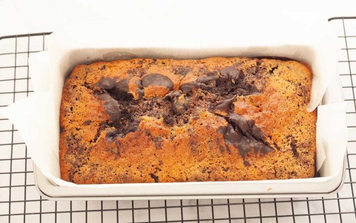 A baked marble loaf cake in a pan lined with parchment paper on a wire rack.