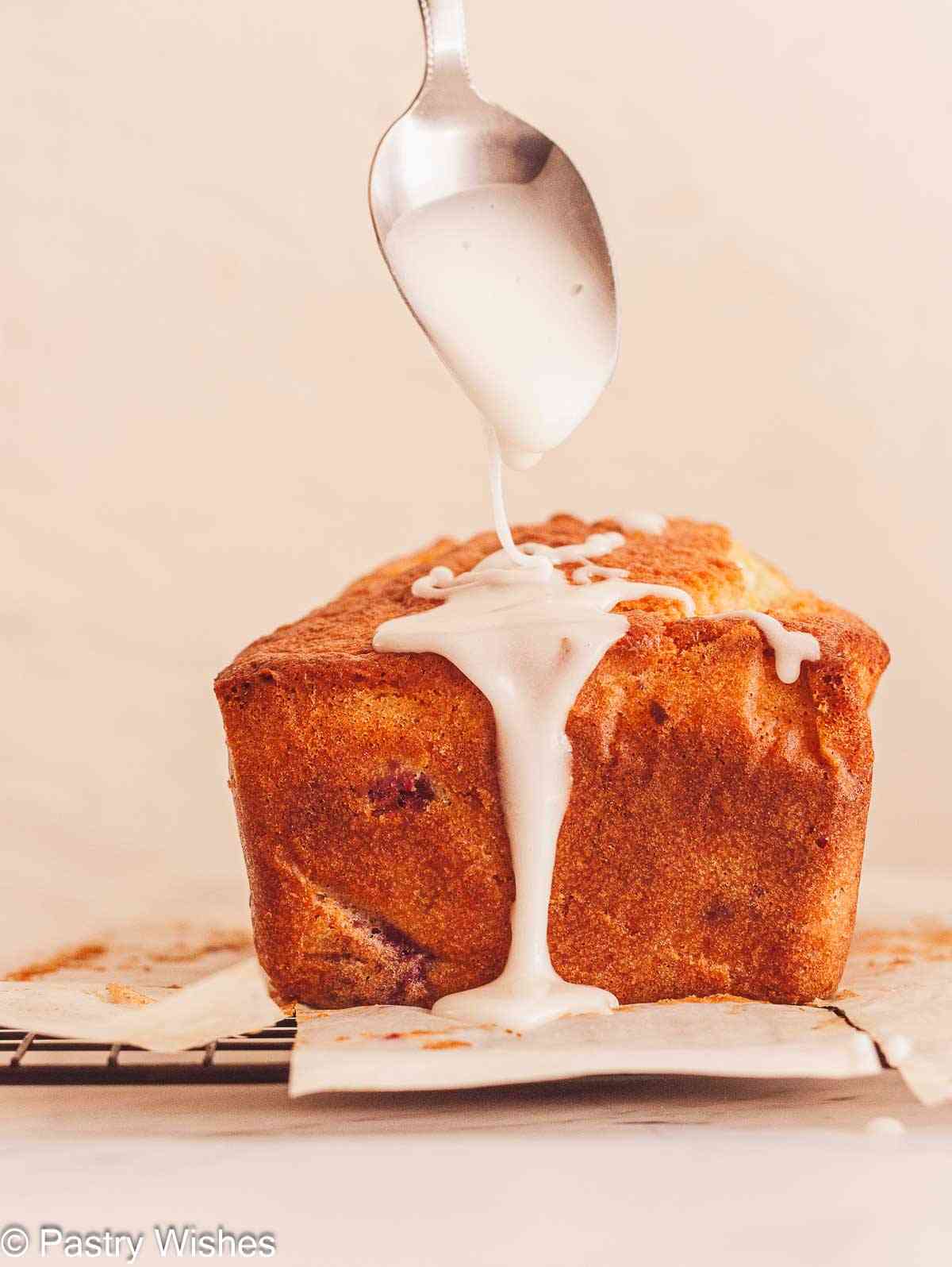 Pouring glaze oveer a lemon raspberry loaf cake with a spoon.