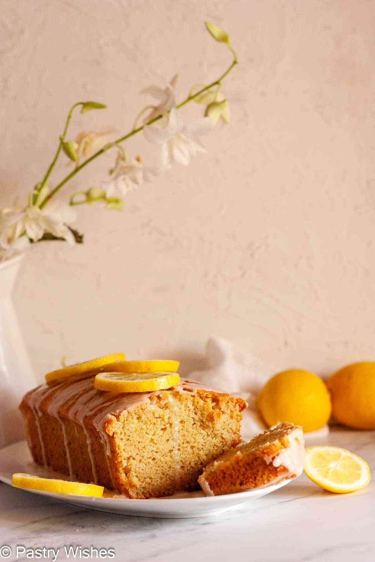 Slice cut from eggless lemon loaf cake with lemon glaze and fresh lemons in the background.