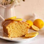 Square image of a sliced eggless lemon loaf cake with lemon glaze and fresh lemons.