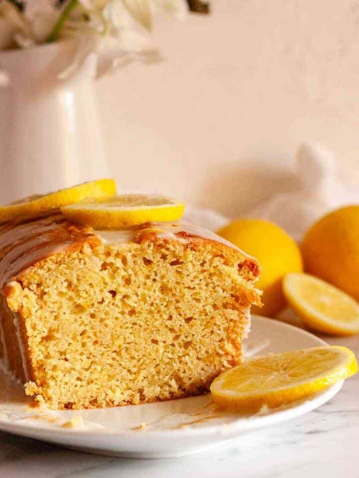 Square image of a sliced eggless lemon loaf cake with lemon glaze and fresh lemons.