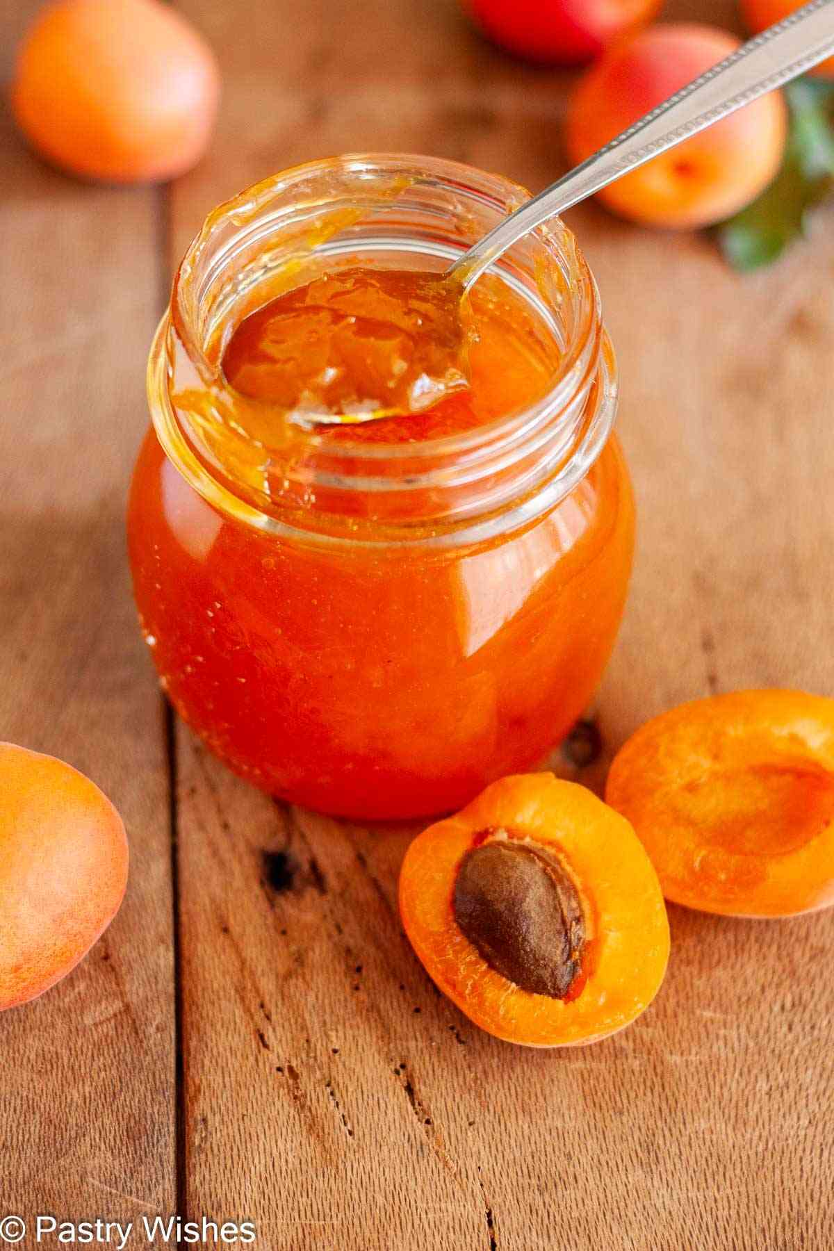 Thick apricot jam on a spoon over a glass jar with fresh apricots on a wooden board.