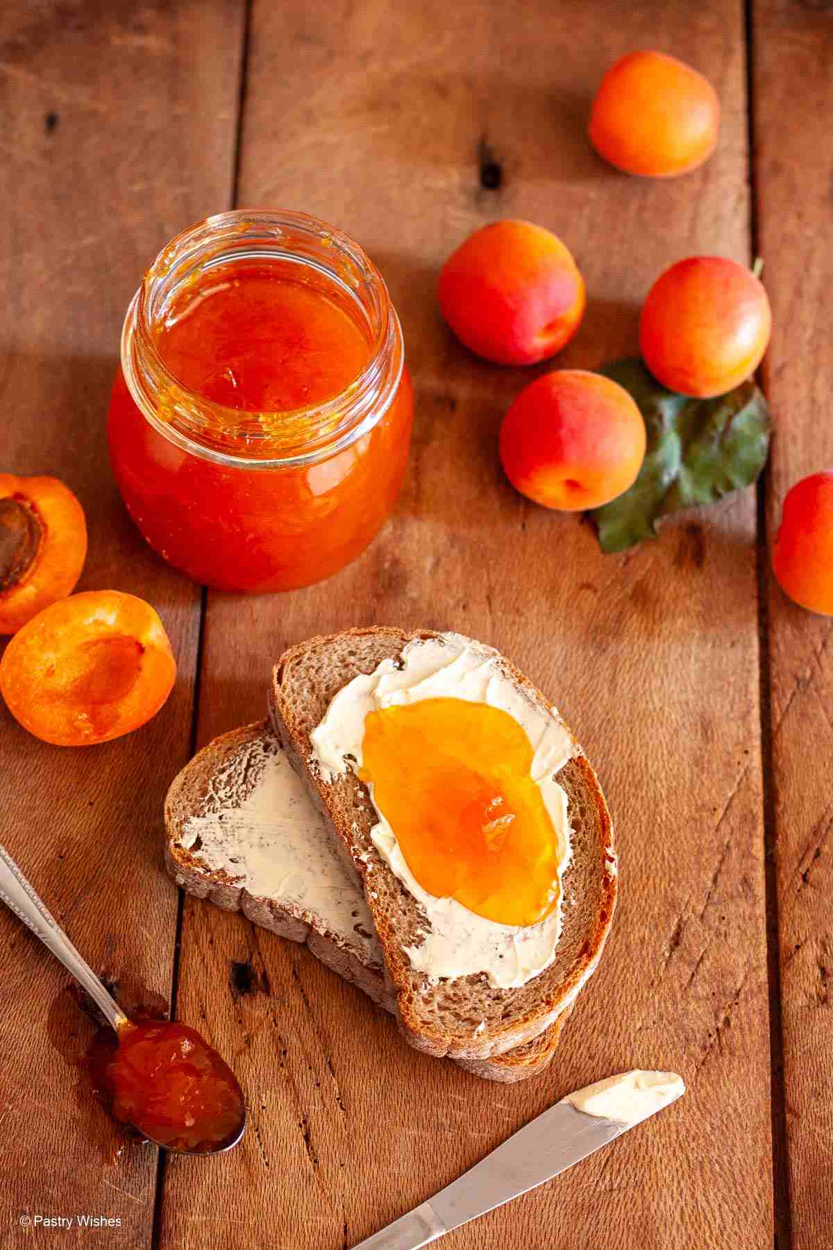 Homemade apricot jam on buttered bread with a jar of jam, fresh apricots, and a spoon on a wooden board.