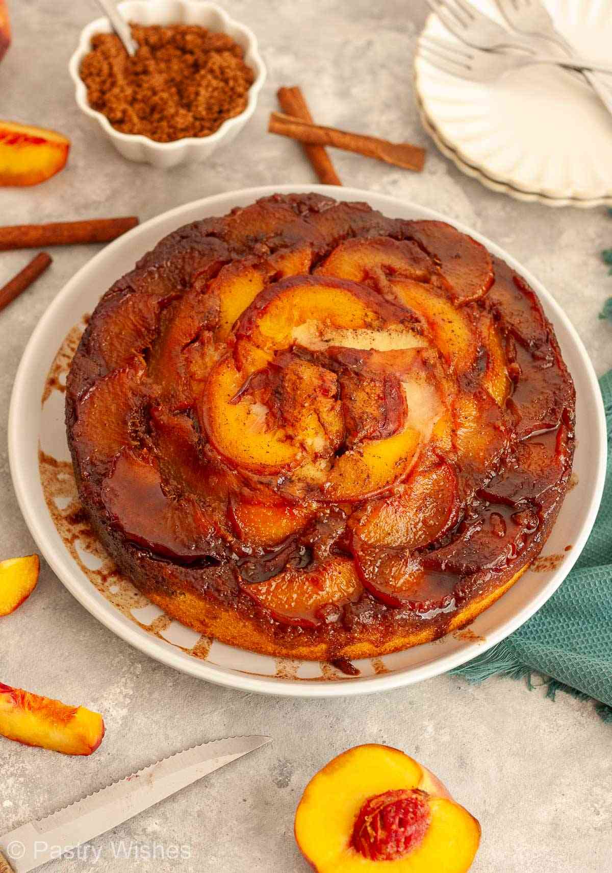 Overhead view of a caramel peach upside-down cake surrounded by fresh peaches and cinnamon sticks.