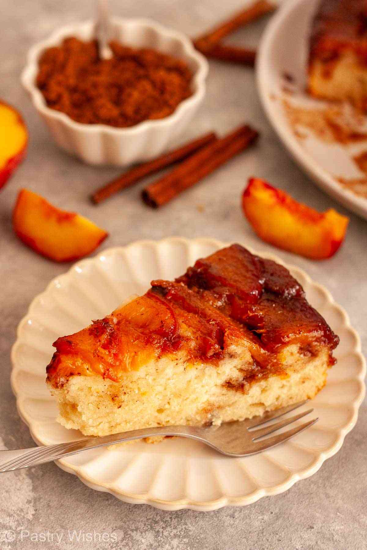 Slice of caramel peach upside-down cake on a scalloped plate with fresh peaches and cinnamon.