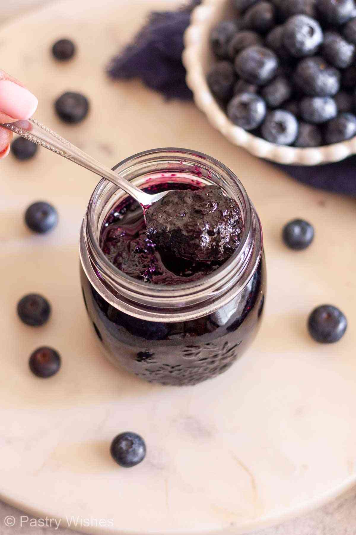 Spoonful of homemade blueberry jam over an open jar with fresh blueberries in the background.