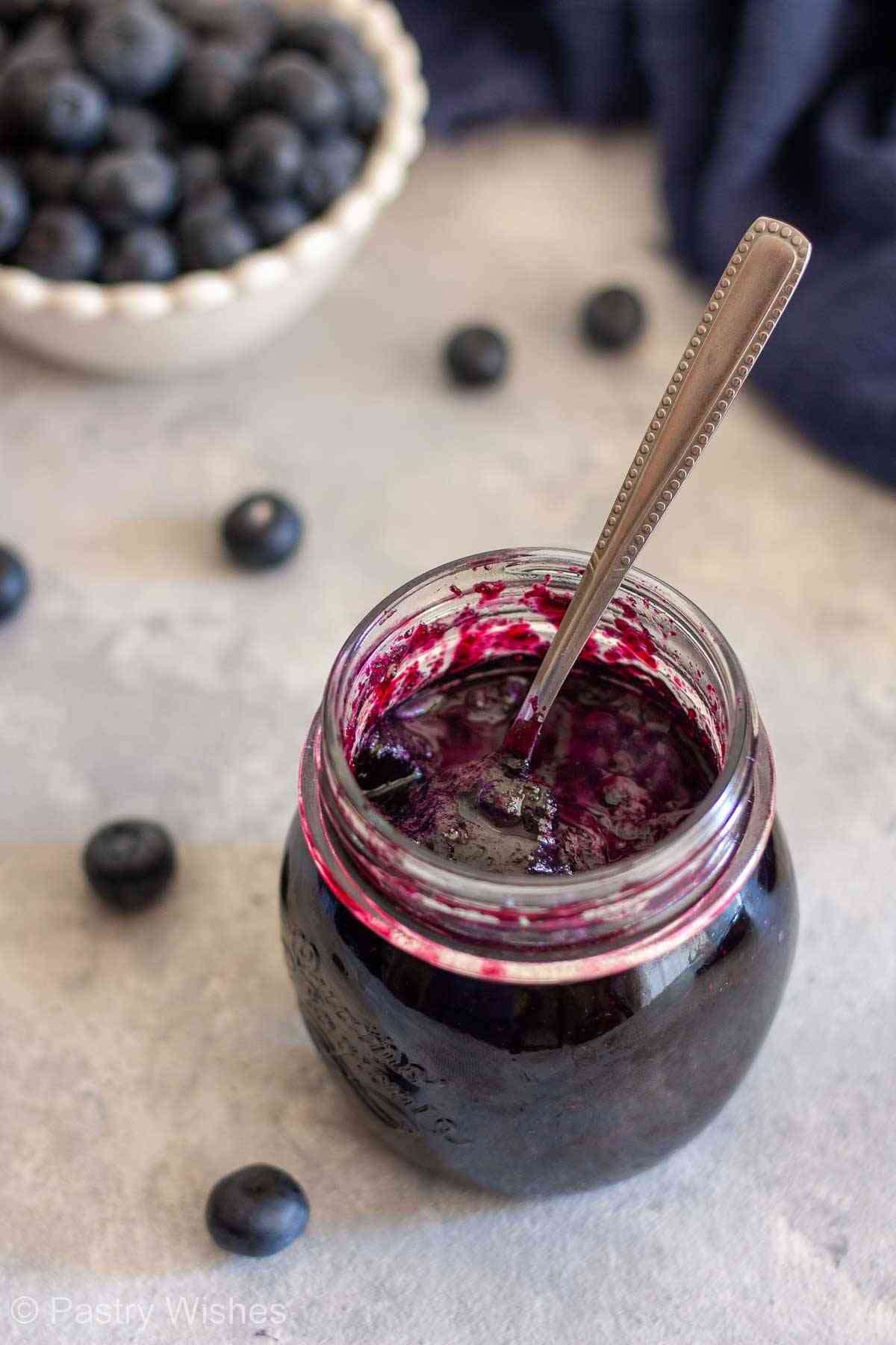 ar of thick homemade blueberry jam with a spoon and fresh blueberries.