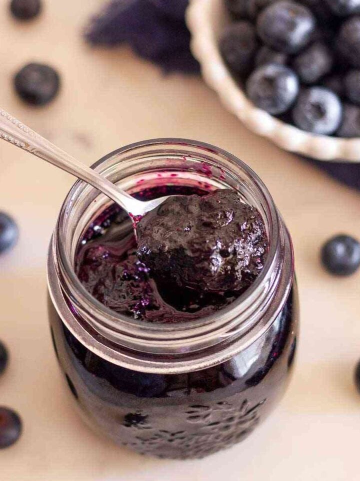 Spoonful of homemade blueberry jam without pectin over an open jar with fresh blueberries in the background.