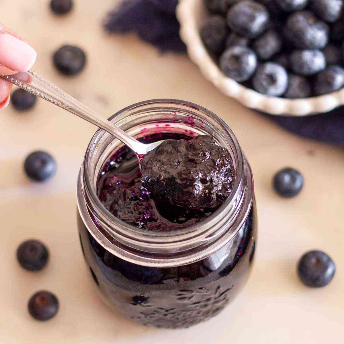 Spoonful of homemade blueberry jam without pectin over an open jar with fresh blueberries in the background.