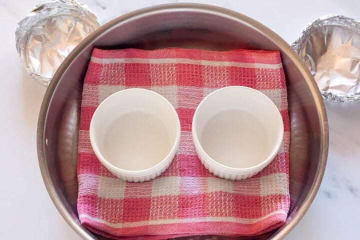 Two empty ramekins on a kitchen towel inside a wide pan, with foil covers beside it.
