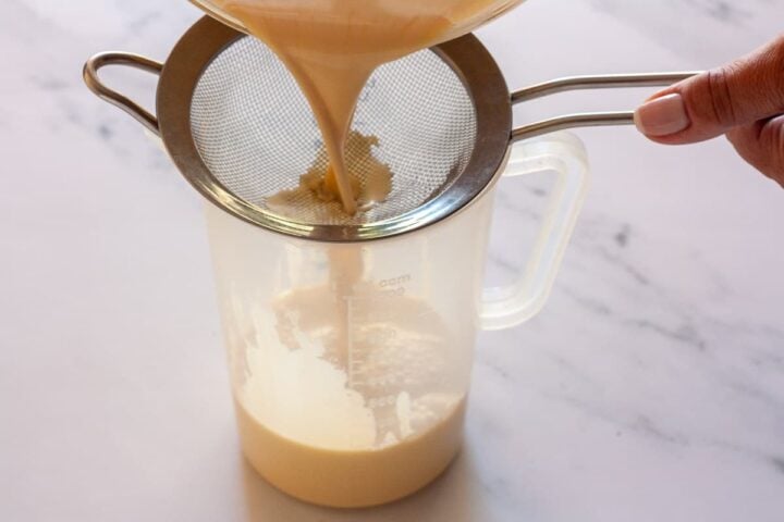 Pouring crème brûlée custard through a fine-mesh sieve into a measuring jug.