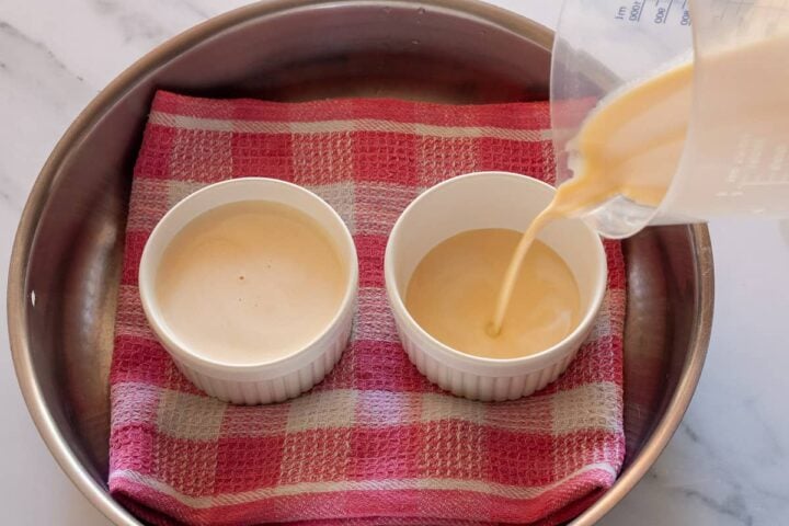 Pouring strained custard into two ramekins in a towel-lined pan.